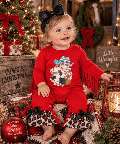 A toddler girl wearing a red long-sleeve romper with fringe, leopard-print ruffle hems, and a Santa graphic on the front, sitting in a rustic Christmas-themed setting with a tree, lantern, and wooden crates.