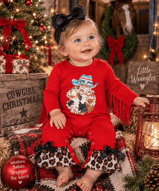 A toddler girl wearing a red long-sleeve romper with fringe, leopard-print ruffle hems, and a Santa graphic on the front, sitting in a rustic Christmas-themed setting with a tree, lantern, and wooden crates.