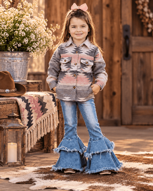 A young girl stands in a rustic barn-like setting wearing a patterned button-front shirt jacket with geometric Southwestern-style stripes in gray, pink, and beige, paired with blue flared jeans that have layered fringe hems. She has a pink bow in her hair, with a wooden door, lantern, blanket, and floral decor in the background.