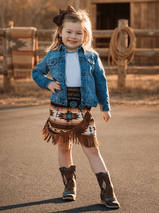 A young girl stands outdoors on a paved road wearing a western-style outfit consisting of a blue denim jacket, a white top, a brown and beige patterned fringed skirt, a turquoise concho belt, and brown cowboy boots. A wooden fence and barn are visible in the background.