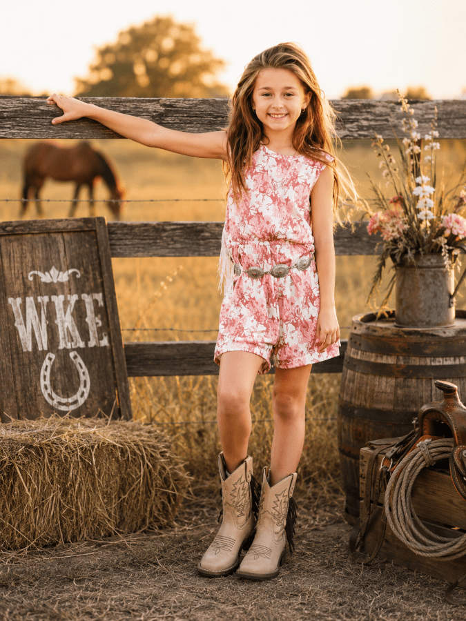 A young girl stands outdoors in a rustic barn-like setting, leaning on a wooden fence. She is wearing a sleeveless pink and white floral romper with a matching belt and beige cowboy boots.