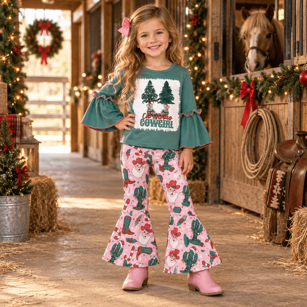 A young girl stands in a rustic barn aisle wearing a festive two-piece outfit. The set includes a teal green long-sleeve top with ruffled cuffs and a front graphic that reads 'Proud COWBOY' beneath Christmas trees, paired with pink bell-bottom pants featuring a cactus, cowboy hat, and Santa pattern.