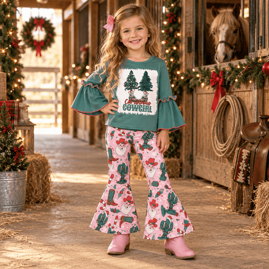 A young girl stands in a rustic barn aisle wearing a festive two-piece outfit. The set includes a teal green long-sleeve top with ruffled cuffs and a front graphic that reads 'Proud COWBOY' beneath Christmas trees, paired with pink bell-bottom pants featuring a cactus, cowboy hat, and Santa pattern.