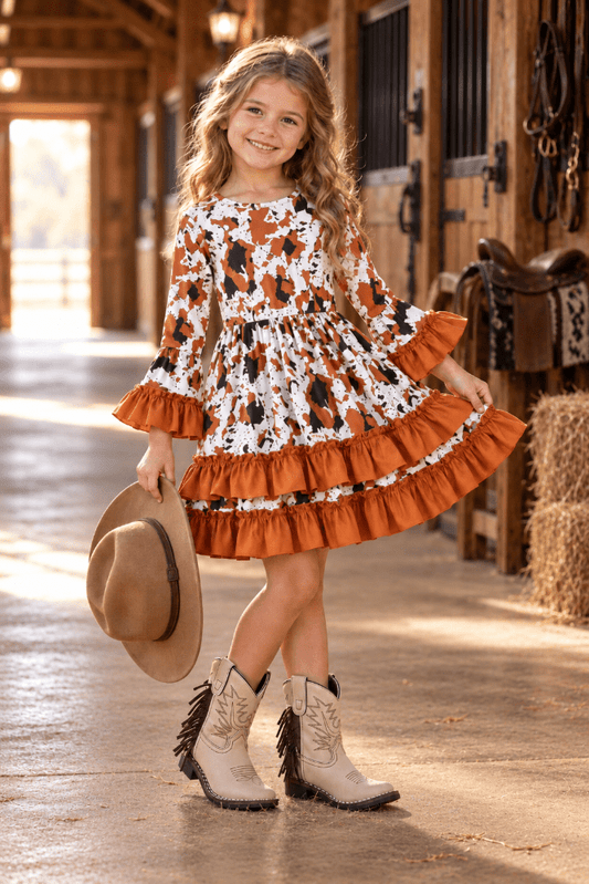 A young girl stands in a rustic barn aisle wearing a tiered long-sleeve dress with an orange, white, and black floral pattern, holding a tan cowboy hat, and wearing beige western boots.