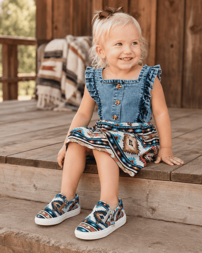 A young child sitting on wooden porch steps wearing a denim overall-style top with a colorful geometric patterned skirt and matching slip-on shoes.