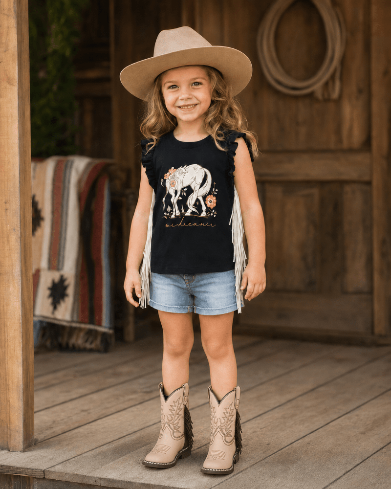 A young girl stands on a wooden porch wearing a black sleeveless fringe top with a white and orange elephant graphic, denim shorts, a tan cowboy hat, and tan cowboy boots.