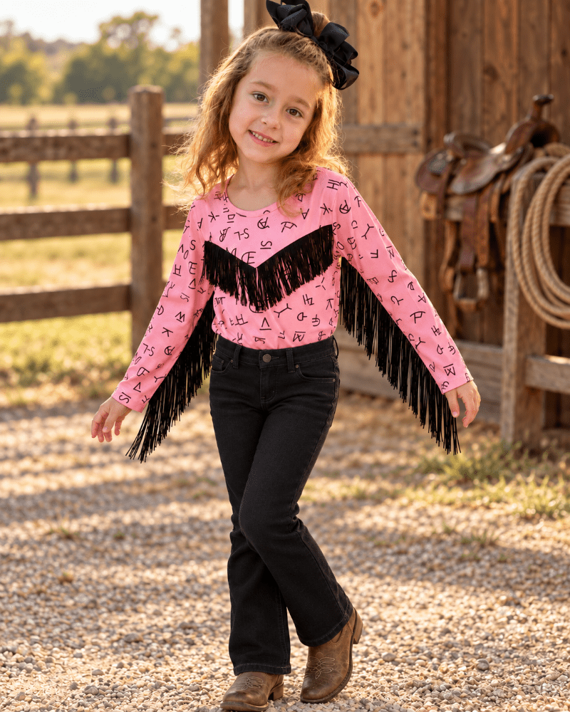 A young girl stands on a gravel path in front of a rustic wooden barn, wearing a pink long-sleeve fringed top with black western-style jeans and brown boots.