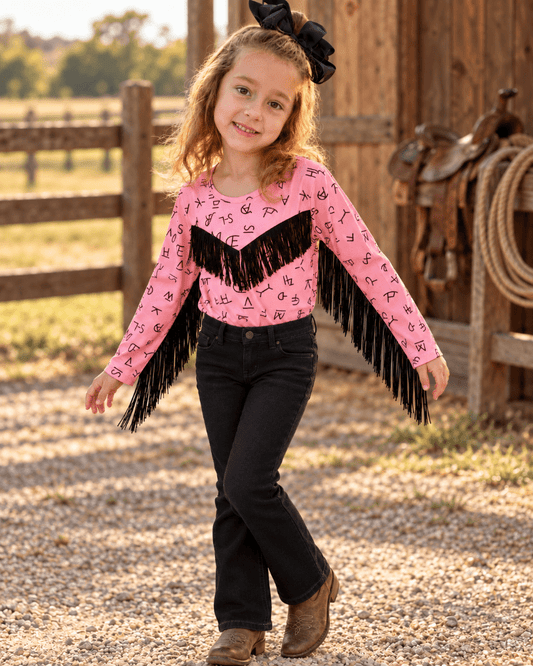 A young girl stands on a gravel path in front of a rustic wooden barn, wearing a pink long-sleeve fringed top with black western-style jeans and brown boots.
