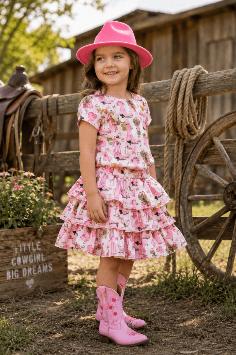 A young girl stands outdoors in a rustic barn setting wearing a pink tiered dress with a cow-print pattern, a bright pink hat, and pink cowboy boots. A wooden fence, rope, wagon wheel, and a sign reading 'LITTLE COWGIRL BIG DREAMS' are visible in the background.