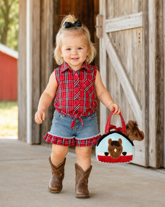 A toddler girl stands in front of a rustic wooden barn door, wearing a red plaid sleeveless button-up top, denim shorts with a plaid ruffle, and brown cowboy boots. She is holding a small red and white felt purse with a cartoon horse design.