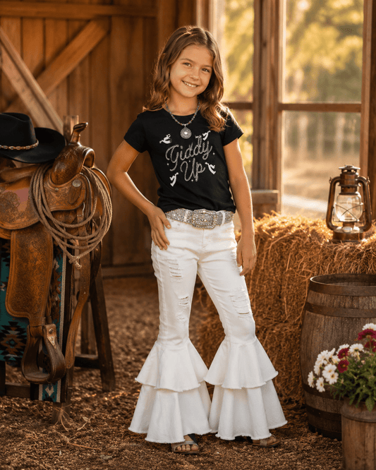 A young girl stands in a rustic barn setting wearing a black graphic t-shirt that reads 'Giddy Up!' and white flared jeans with ruffled tiers. The background features a wooden saddle, hay bale, lantern, and barrel.