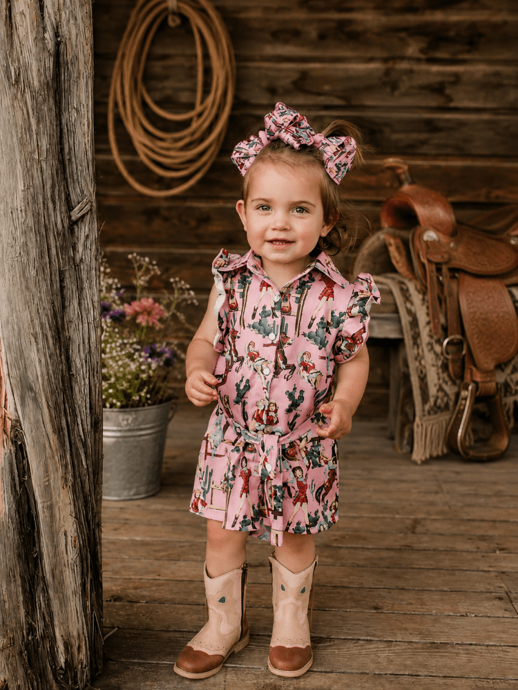 A toddler girl stands on a rustic wooden porch wearing a pink western-style printed romper with a matching bow headband and cowboy boots. A wooden log, rope coil, and saddle are visible in the background.
