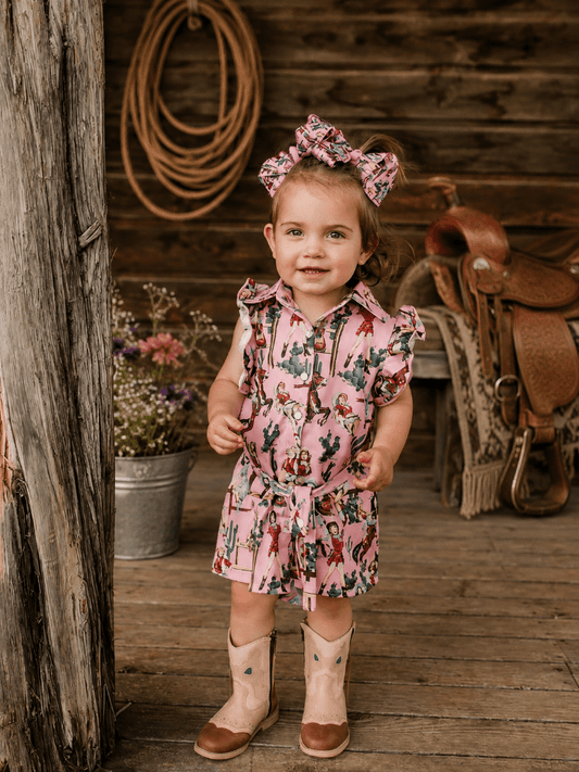 A toddler girl stands on a rustic wooden porch wearing a pink western-style printed romper with a matching bow headband and cowboy boots. A wooden log, rope coil, and saddle are visible in the background.
