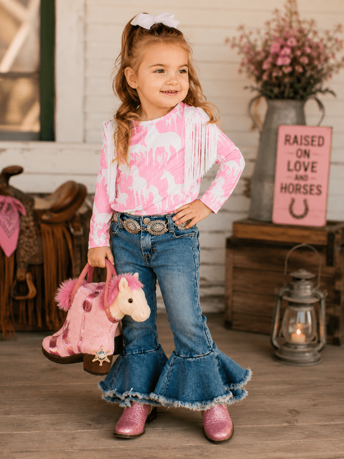 A young girl stands in a rustic western-themed room wearing a pink and white patterned long-sleeve top with fringe, blue flared jeans, and pink boots. She is holding a pink plush horse toy bag.
