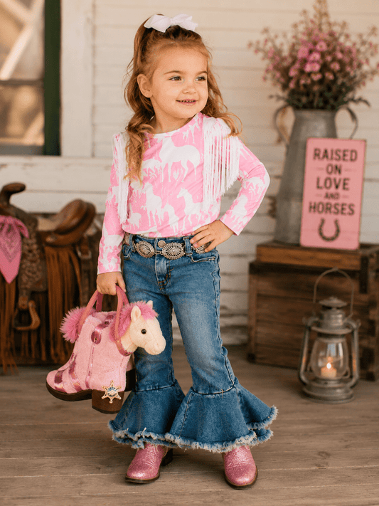 A young girl stands in a rustic western-themed room wearing a pink and white patterned long-sleeve top with fringe, blue flared jeans, and pink boots. She is holding a pink plush horse toy bag.