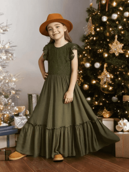 Young girl in a green dress and brown hat standing in front of a decorated Christmas tree.