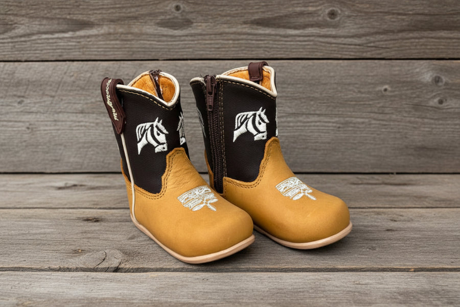 Pair of tan cowboy boots with brown accents and white horse designs on a light wooden background