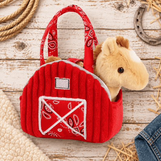 A red plush horse-shaped handbag with a white bandana barn emblem on the front, shown on a rustic whitewashed wood surface with rope, a horseshoe, straw, and denim props.