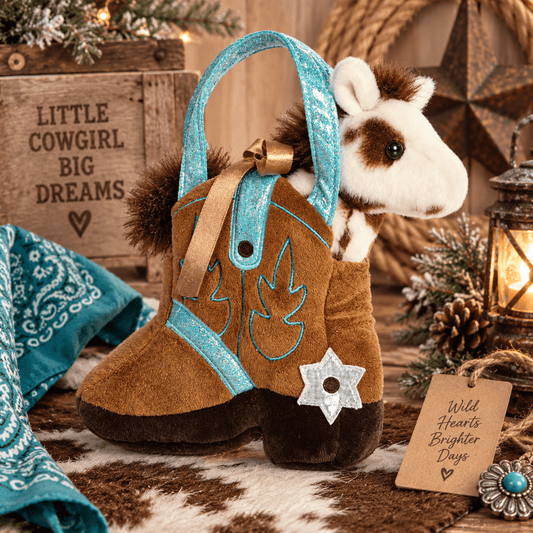 A western-themed plush toy featuring a brown cowboy boot with turquoise glitter trim and a small plush horse peeking out from behind the side. The toy is styled on a cowhide rug with a turquoise bandana, a wooden sign reading 'Little Cowgirl Big Dreams,' and warm holiday decorations in the background.