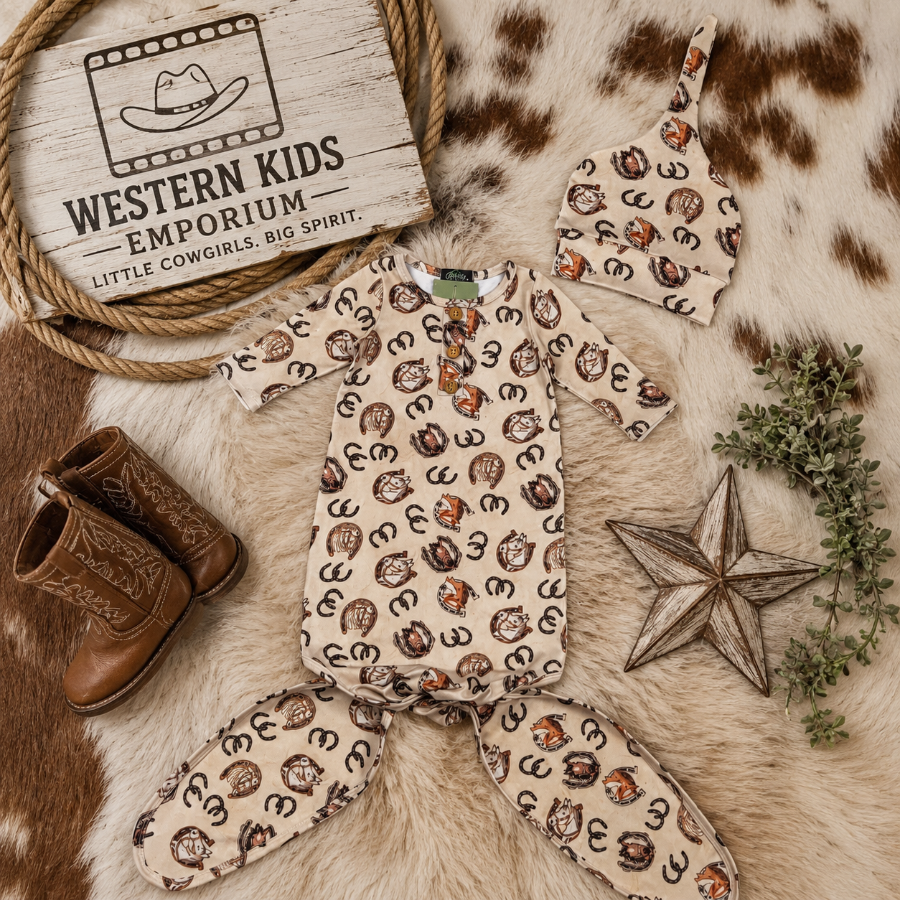 A baby western-themed clothing set from Western Kids Emporium, featuring a short-sleeve romper and matching footed pants in a beige print with brown and black horse silhouettes, styled on a cowhide rug with cowboy boots, a wooden star, greenery, and a branded sign.