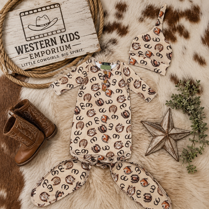 A baby western-themed clothing set from Western Kids Emporium, featuring a short-sleeve romper and matching footed pants in a beige print with brown and black horse silhouettes, styled on a cowhide rug with cowboy boots, a wooden star, greenery, and a branded sign.