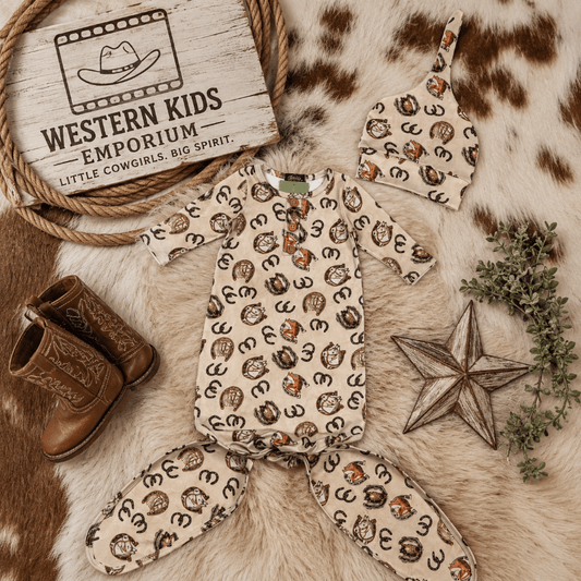 A baby western-themed clothing set from Western Kids Emporium, featuring a short-sleeve romper and matching footed pants in a beige print with brown and black horse silhouettes, styled on a cowhide rug with cowboy boots, a wooden star, greenery, and a branded sign.