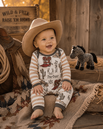 Baby in cowboy outfit with hat and western-themed clothing, surrounded by rustic decor.