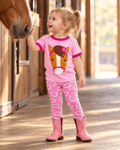 A young girl in a pink ringer t-shirt with a horse face graphic and matching pink horseshoe-print leggings stands in a stable aisle, reaching out to a brown horse's nose while wearing pink cowboy boots.
