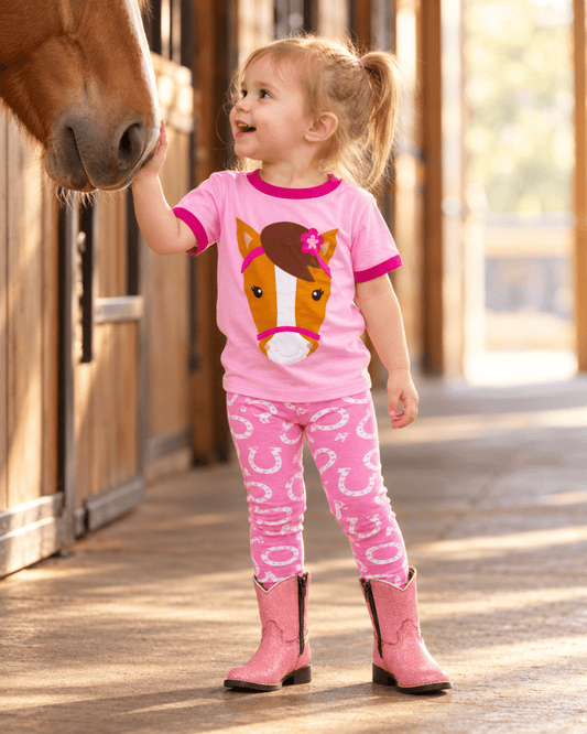 A young girl in a pink ringer t-shirt with a horse face graphic and matching pink horseshoe-print leggings stands in a stable aisle, reaching out to a brown horse's nose while wearing pink cowboy boots.
