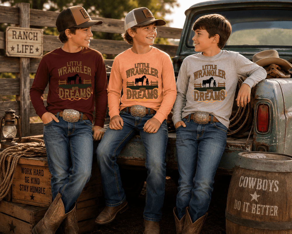 Three boys wearing matching 'Little Wrangler Dreams' long-sleeve cowboy shirts, jeans, and boots, posing together in a rustic outdoor setting with a vintage pickup truck, wooden fence, and barrels.