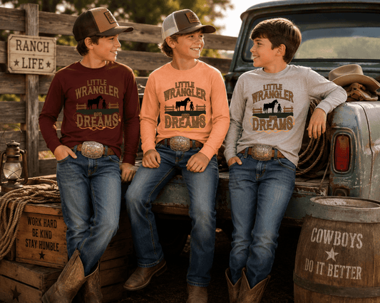 Three boys wearing matching 'Little Wrangler Dreams' long-sleeve cowboy shirts, jeans, and boots, posing together in a rustic outdoor setting with a vintage pickup truck, wooden fence, and barrels.