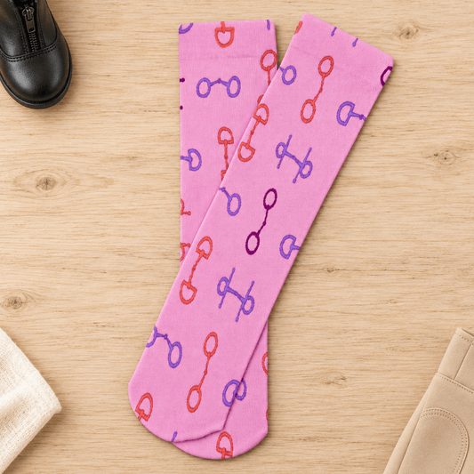 A pair of pink knee-high socks with an allover equestrian horse-bit pattern in red, purple, and blue, laid flat on a light wood background with styled equestrian accessories.