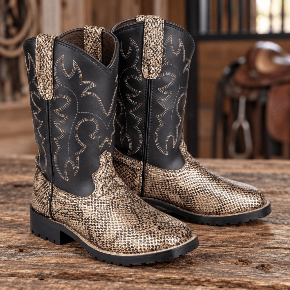 A pair of western cowboy boots with a black shaft and a metallic gold snakeskin-patterned foot, displayed on a rustic wooden table with a blurred saddle in the background.