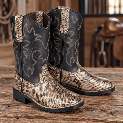 A pair of western cowboy boots with a black shaft and a metallic gold snakeskin-patterned foot, displayed on a rustic wooden table with a blurred saddle in the background.