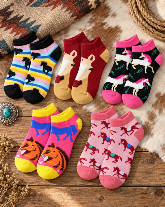 Six pairs of children's ankle socks arranged in a flat lay on a rustic wooden surface with western-style props. The socks feature colorful horse and unicorn patterns in pink, red, yellow, blue, black, and white.