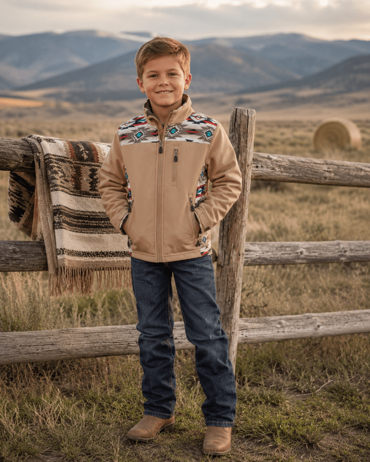 A young boy stands outdoors in a rural landscape wearing a tan zip-front jacket with colorful geometric patterned yoke panels, blue jeans, and brown boots. A rustic wooden fence and a patterned throw blanket are visible behind him, with mountains in the background.