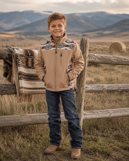 A young boy stands outdoors in a rural landscape wearing a tan zip-front jacket with colorful geometric patterned yoke panels, blue jeans, and brown boots. A rustic wooden fence and a patterned throw blanket are visible behind him, with mountains in the background.