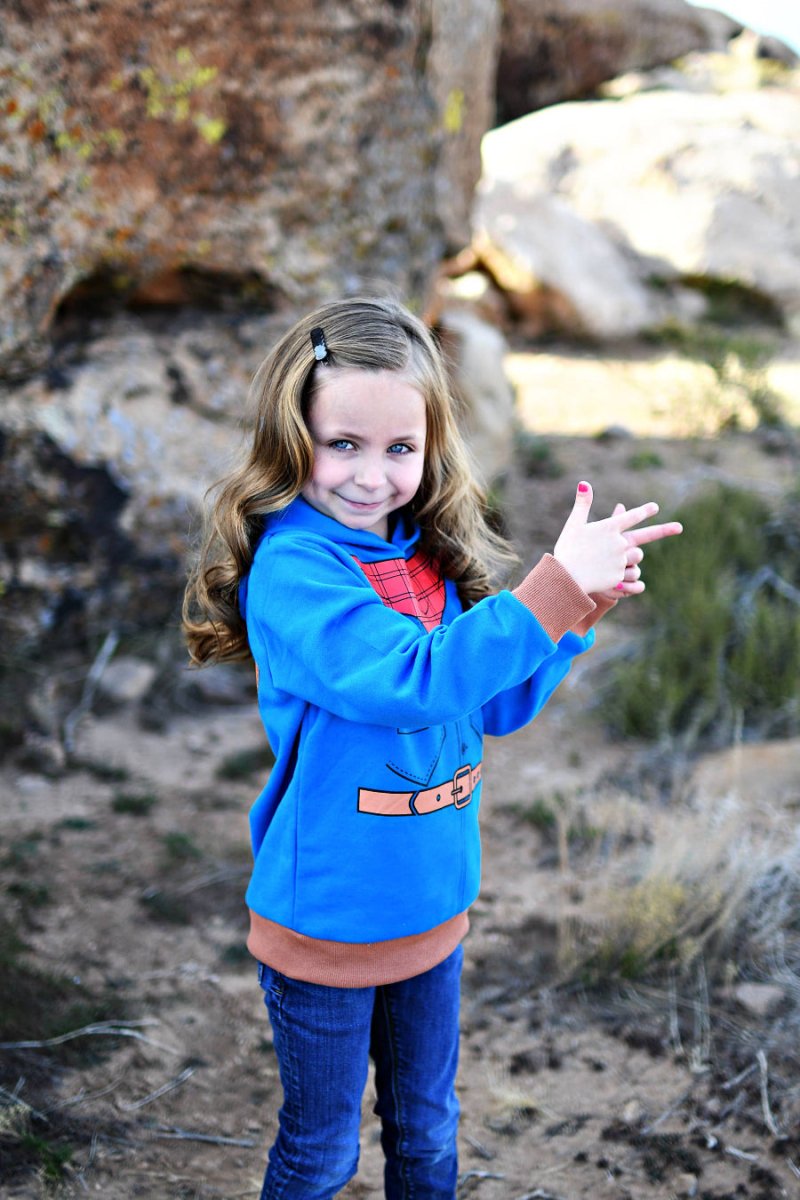 Child in a blue hoodie with a bandana standing in a rocky outdoor setting.