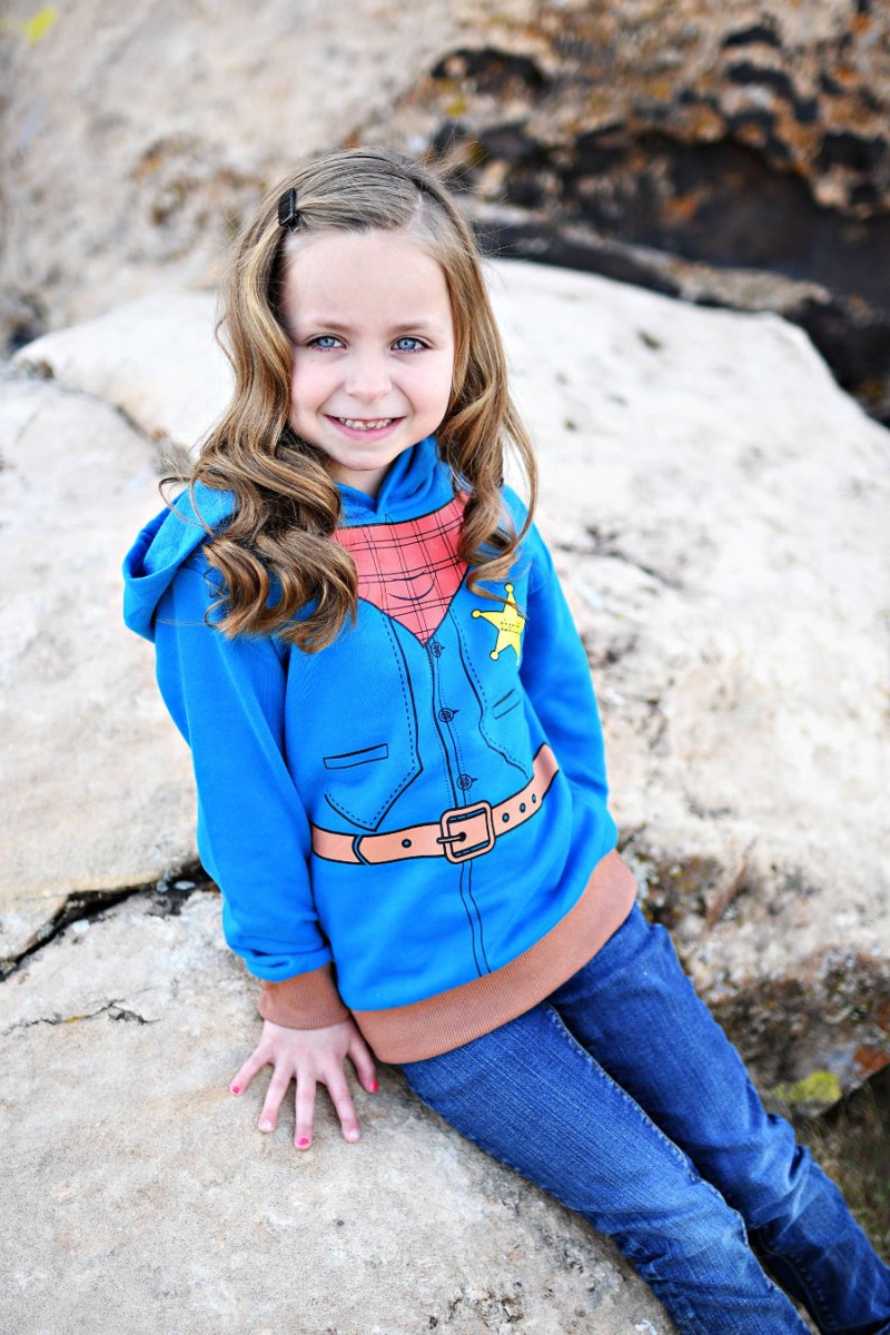 Young girl in a blue hoodie with cowboy design sitting on rocks.