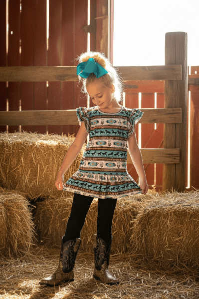 Young girl in a patterned pants set and cowboy boots standing in a barn with hay bales.