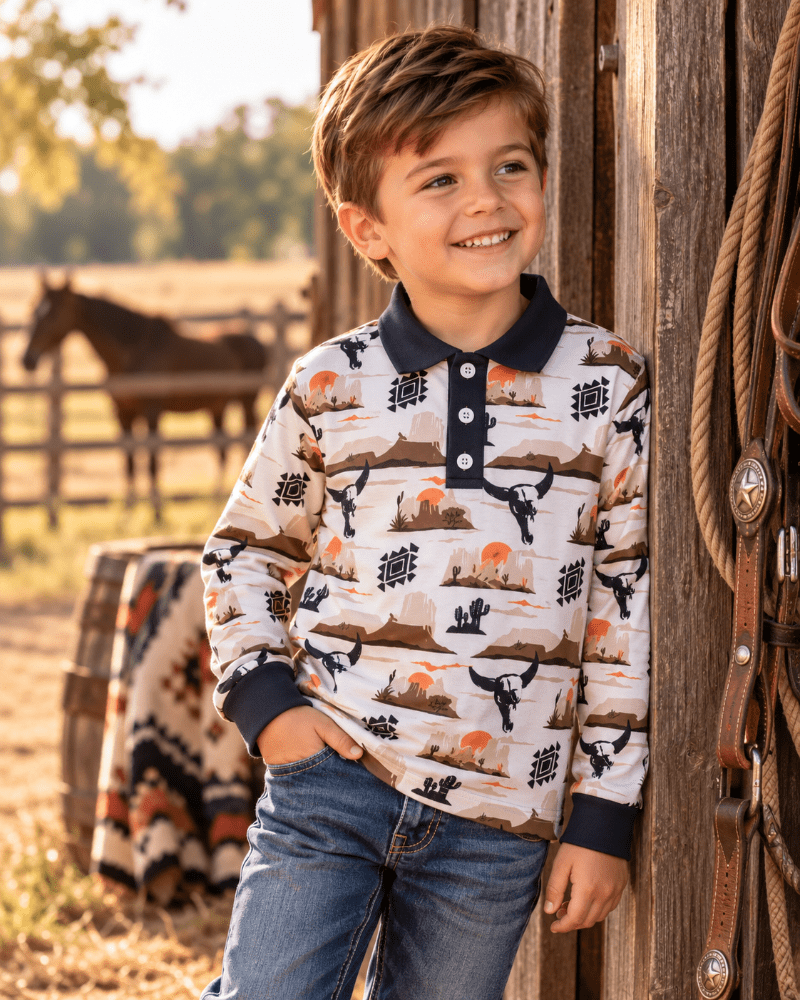 A young boy smiling and looking to the side, wearing a long-sleeve polo shirt with a western desert landscape print and blue jeans, standing next to a rustic wooden barn wall with a horse in the background.