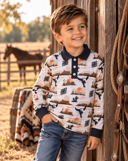 A young boy smiling and looking to the side, wearing a long-sleeve polo shirt with a western desert landscape print and blue jeans, standing next to a rustic wooden barn wall with a horse in the background.