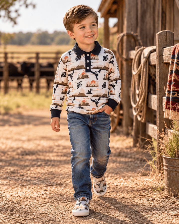 A young boy walking outdoors on a dirt path next to a rustic wooden fence and barn, wearing a long-sleeve polo shirt with a western tree pattern, blue jeans, and sneakers.