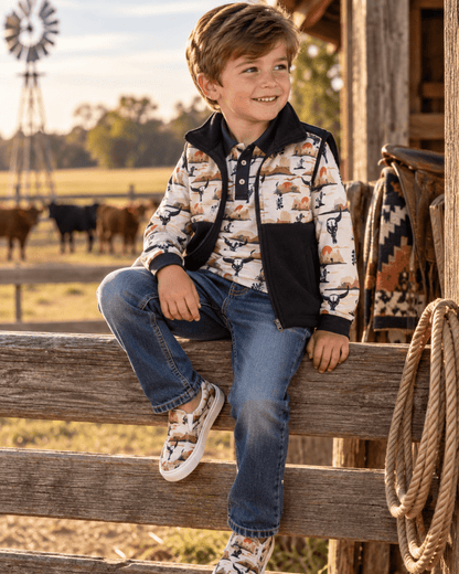 A young boy wearing a western-style patterned jacket, blue jeans, and matching patterned sneakers sits on a wooden fence in a rustic barn yard with a windmill and cows in the background.