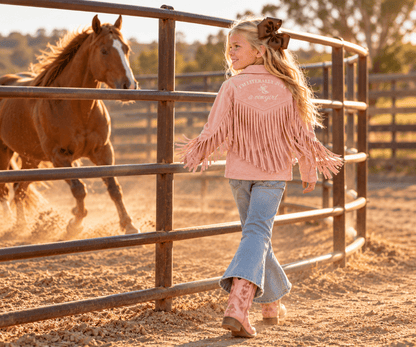 A young girl wearing a pink fringed western-style jacket, blue jeans, and pink boots walks beside a metal fence in a dusty ranch paddock, with a brown horse running in the background.