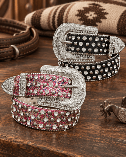 Two western-style studded belts displayed on a rustic wooden table, one with a silver buckle and pink glitter body, and the other with a silver buckle and black glitter body. A spurs and rolled blanket are visible in the background.