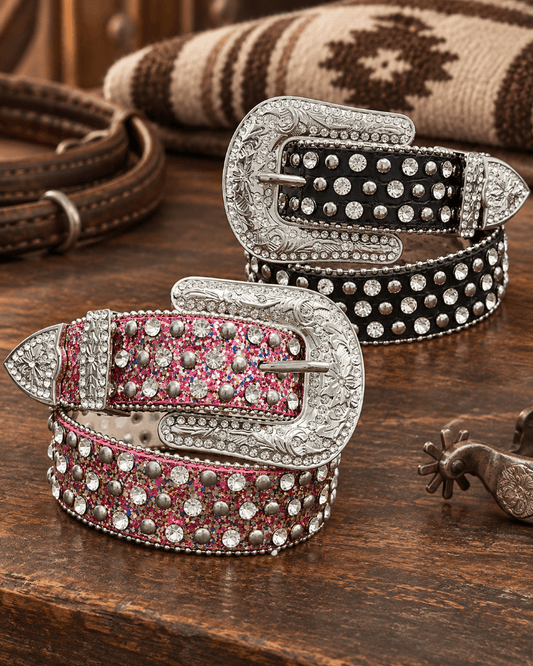 Two western-style studded belts displayed on a rustic wooden table, one with a silver buckle and pink glitter body, and the other with a silver buckle and black glitter body. A spurs and rolled blanket are visible in the background.