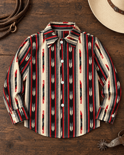 A long-sleeve western-style button-up shirt with a Southwestern geometric pattern in red, black, cream, and white, laid flat on a rustic wooden surface with a cowboy hat, leather strap, and metal spur as styling props.