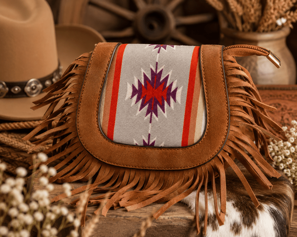 A brown suede fringe bag with a Southwestern-style woven panel in gray, red, and purple, styled on a rustic wooden surface with a cowboy hat and dried flowers.