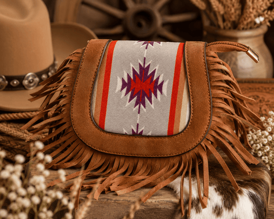 A brown suede fringe bag with a Southwestern-style woven panel in gray, red, and purple, styled on a rustic wooden surface with a cowboy hat and dried flowers.